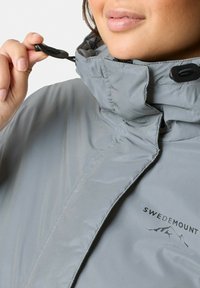 Grijze geïsoleerde jas met een hoge kraag, verstelbare trekkoord en het logo "SWEDEMOUNT" aan de voorkant. Gladde textuur en waterdicht materiaal.