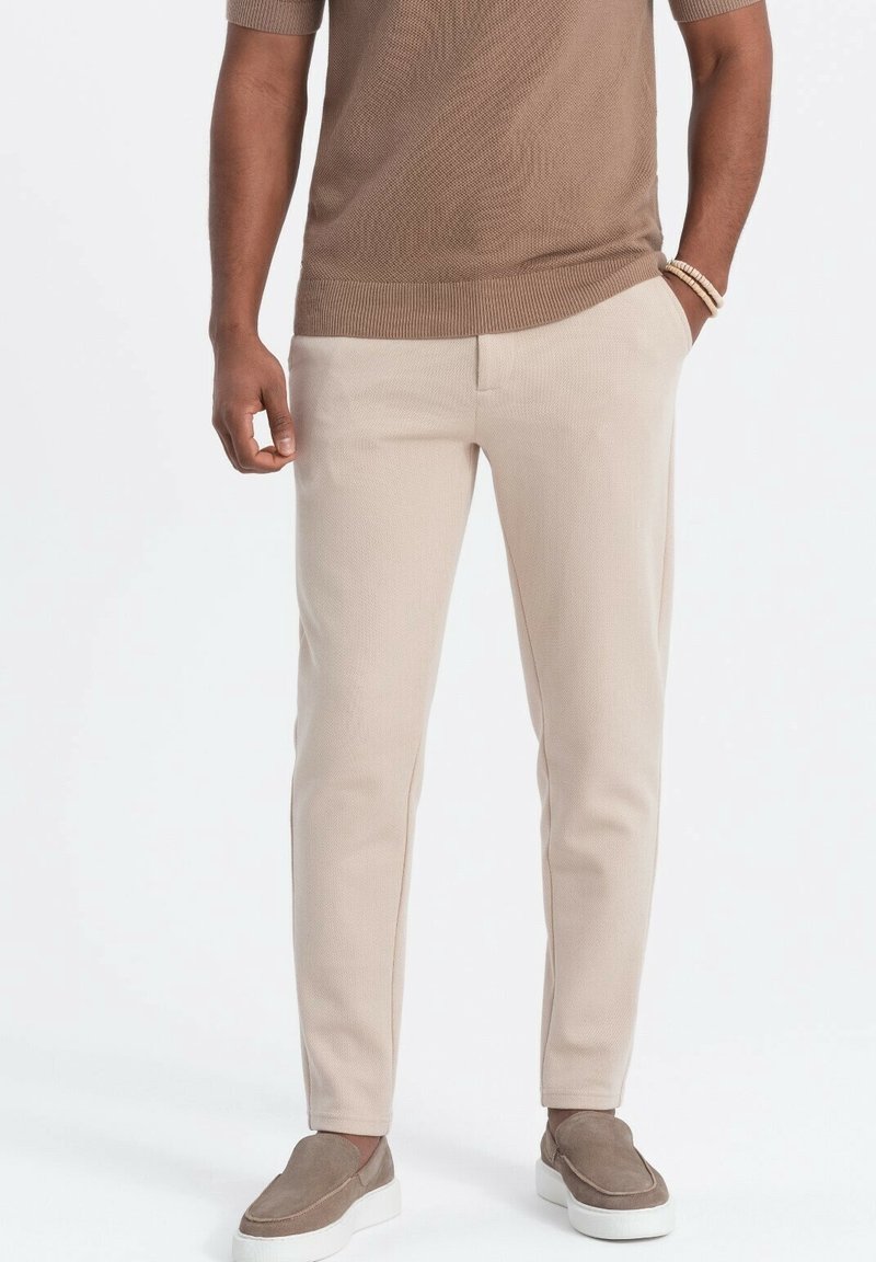 Pantalon beige ajusté en tissu texturé, avec poches latérales et une ceinture lisse, associé à des chaussures grises sans lacets.