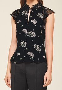 Sort ærmeløs bluse med gennemsigtige rufflede ærmer, blomstermotiv og en lille nøglehulsudskæring, båret med sorte bukser.