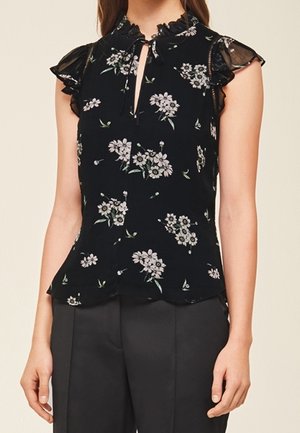 Blouse noire sans manches avec des manches chauve-souris transparentes et volantées, imprimé floral et un petit décolleté en trou de serrure, portée avec un pantalon noir.