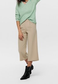 Pantalon beige à jambes larges et taille haute, associé à un pull en maille vert menthe et des bottines noires. Texture lisse, coupe décontractée.