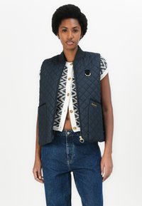 Colete acolchoado azul marinho com gola alta, bolsos frontais e detalhes em metal dourado, sobre um suéter estampado e jeans de ganga.