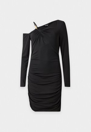 Une femme porte une robe noire froncée avec une découpe au décolleté, un sac à main bordeaux et des talons noirs.