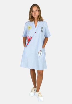 Woman wearing light blue short-sleeve dress with embroidered lobster, sun, sunglasses, and sandals, standing with hands in pockets and white sneakers.