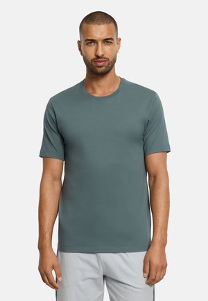 Mann mit kurzen Haaren und Bart, der ein einfarbiges türkisfarbenes T-Shirt mit Rundhalsausschnitt und kurzen Ärmeln sowie hellgraue Shorts trägt, steht vor einem weißen Hintergrund.