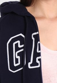 Sweat à capuche bleu marine avec les lettres "GA" brodées en blanc. Doté d'une fermeture éclair à l'avant et d'un tissu doux et texturé. Chemise rose clair visible en dessous.