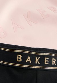 Zwarte stof met een textuurband heeft "BAKER" in gouden letters, tegen een zachte roze achtergrond met reliëftekst "BAKER".