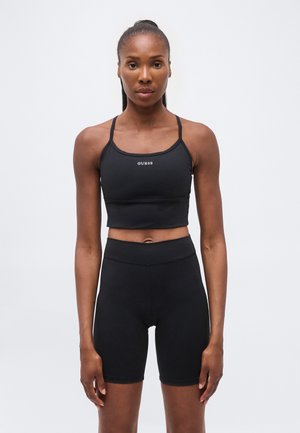 Atletische vrouw die naar voren kijkt, draagt een zwarte Guess crop top en zwarte high waist biker shorts tegen een effen achtergrond.