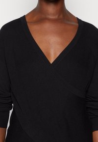 Schwarzer Wickelpullover aus strukturiertem Strickstoff. V-Ausschnitt-Design mit langen Ärmeln und diagonalen Nahtdetails an der Vorderseite.