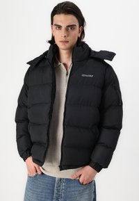 Μαύρο μπουφάν πούφφας με ψηλό γιακά, τσέπες με φερμουάρ, ελαστικούς μανσέτες και λογότυπο στο στήθος, φορεμένο πάνω από ένα ανοιχτό μπεζ πουκάμισο.