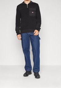 Schwarzer Strickpullover mit einem Frontreißverschluss, ausgestattet mit einer Tasche mit Logo-Detail. Kombiniert mit lockeren blauen Jeans und schwarzem Schuhwerk.