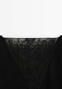 Encolure de vêtement noire avec dentelle transparente et petits détails de perles, avec une étiquette de marque cousue à l'intérieur du col.