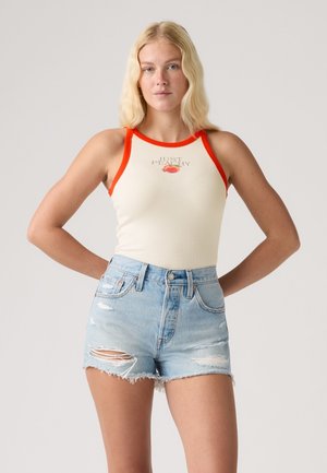 Jeune femme aux cheveux blonds portant un débardeur crème avec une bordure orange et le texte « Just Peachy », associé à un short en jean bleu clair déchiré.
