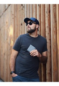 Gray wallet held in a man's hand, wearing a dark shirt and navy cap, sunglasses, against a wooden fence background.