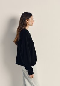 Črna pletena majica z ohlapnim krojem, z dolgimi rokavi in ribanim obodom, v kombinaciji s svetlo modrimi kavbojkami, prikazana s strani.