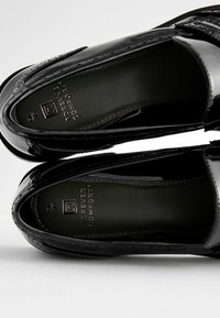 Mocasines de charol negro con un acabado suave, puntera redondeada y logo en relieve en la plantilla. Presentan detalles de costura sutiles.