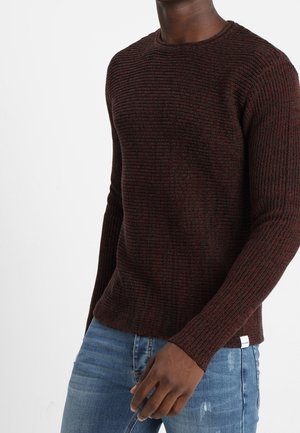 Pull en maille bordeaux avec un design texturé et des manches longues. Doté d'un col rond et d'une petite étiquette sur le côté inférieur. Associé à un jean bleu.