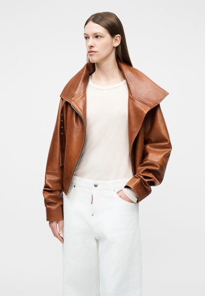 Νεαρή γυναίκα που φορά καφέ δερμάτινο μπουφάν πάνω από λευκό πουκάμισο και άνετο λευκό παντελόνι, στέκεται με το ένα χέρι στην τσέπη, κοιτάζοντας προς τα πλάγια.