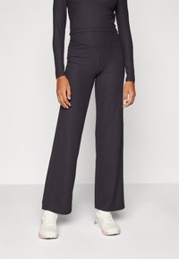 Sort ribbet langærmet top parret med brede sorte bukser. Modellen har lyse farvede sportssko på. Stoffet har en tekstureret finish.