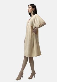 Cremefarbenes, strukturiertes Kleid mit tiefem V-Ausschnitt und kurzen Ärmeln, das einen lockeren Schnitt und seitliche Taschen aufweist. Kombiniert mit nude-farbenen High Heels.
