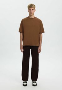 Bruine T-shirt met korte mouwen en een ontspannen pasvorm, gecombineerd met donkerbruin wijde broek en zwarte schoenen met witte accenten. Model staat rechtop.
