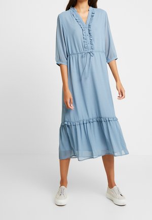 Vrouw draagt een lichtblauwe midi-jurk met ruches, driekwartmouwen en witte sneakers, op een effen achtergrond.