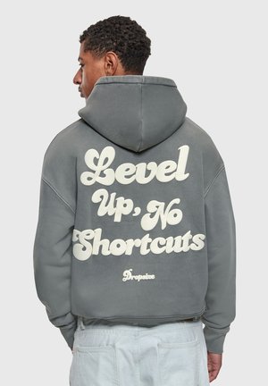 Mężczyzna stojący bokiem, ubrany w szarą bluzę z kapturem z napisem "Level Up, No Shortcuts" oraz białe spodnie na jednolitym tle.