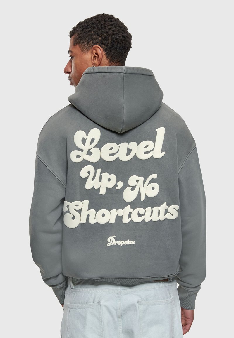 Homme de profil portant un sweat à capuche gris avec le texte « Level Up, No Shortcuts » et un pantalon blanc sur un fond uni.