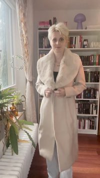 Person med kort blondt hår som knapper igjen en beige frakk med pelskrage, står i et rom med bokhyller, planter og et vindu.