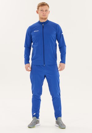 Mand iført en blå zip-up jakke og matchende bukser med grå sportssko, stående mod en ensfarvet hvid baggrund.