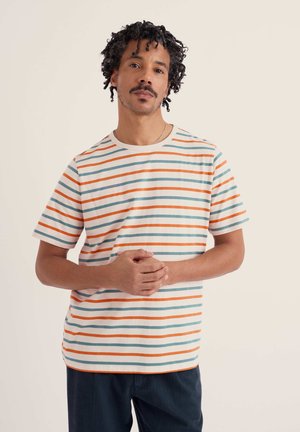 Lühikeste lokkis juustega mees, kellel on valge T-särk oranžide ja siniste horisontaalsete triipudega, käed kokkusirutatud, seisab ühevärvilise tausta ees.