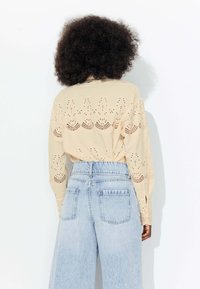 Chemise beige à manches longues avec des détails en broderie à œillets. Associée à un jean en denim bleu clair taille haute. Texture douce, design féminin.