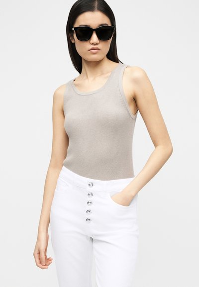 Γυναίκα με μαύρα γυαλιά ηλίου, αμάνικο μπεζ ριμπ τοπ και ψηλόμεσο άσπρο παντελόνι με έξι κουμπιά στο μπροστινό μέρος, ποζάρει με το ένα χέρι στην τσέπη.