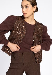 Vrouw die een bruin paillettenvest over een fuzzy bruine trui aanpast, in een bruine broek, met haar in een lage bun en gouden creolen.