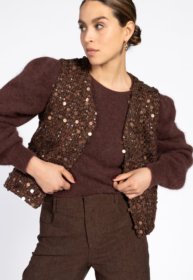 Vrouw die een bruin paillettenvest over een fuzzy bruine trui aanpast, in een bruine broek, met haar in een lage bun en gouden creolen.