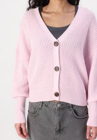Růžový pletený kardigan s výstřihem do V, třemi velkými hnědými knoflíky a žebrovanými manžetami. Nositelný přes šedý top, s volným a krátkým střihem.