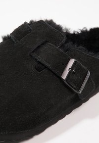 Zoccoli neri in suede con un morbido rivestimento in pelliccia, una fascia larga con una fibbia metallica e una suola in gomma testurizzata.
