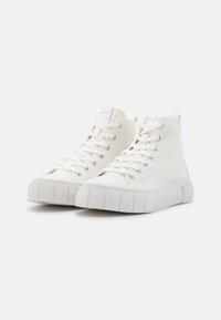 Witte hoge canvas sneakers met dikke rubberen zolen en witte veters, naast elkaar geplaatst op een eenvoudige witte achtergrond.