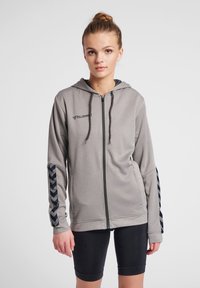 Grauer Reißverschluss-Hoodie mit Kapuze, aus strukturiertem Stoff und schwarzen Chevron-Akzenten an den Ärmeln. Logo auf der oberen linken Seite.