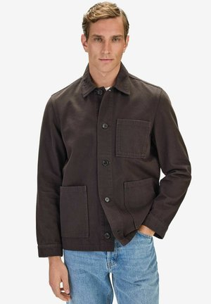 Man som bär en mörkbrun jacka med knappar och tre framfickor samt ljusblå jeans, stående med ena handen i fickan.
