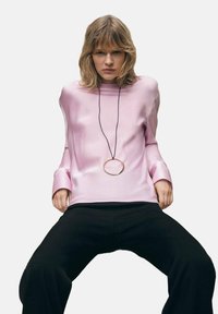 Top lungo a maniche corte in raso rosa con collo alto e polsini ampi. Presenta un grande pendente circolare appeso a un cordino nero. Abbinato a pantaloni neri.