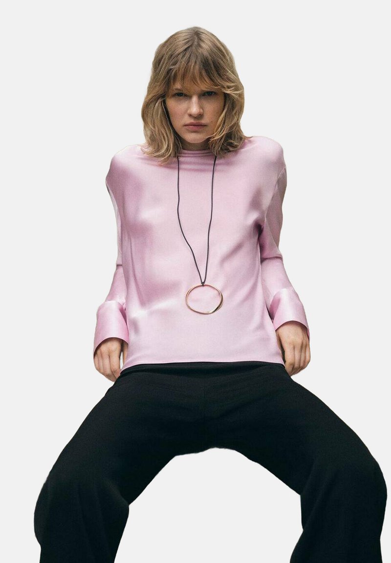 Top lungo a maniche corte in raso rosa con collo alto e polsini ampi. Presenta un grande pendente circolare appeso a un cordino nero. Abbinato a pantaloni neri.