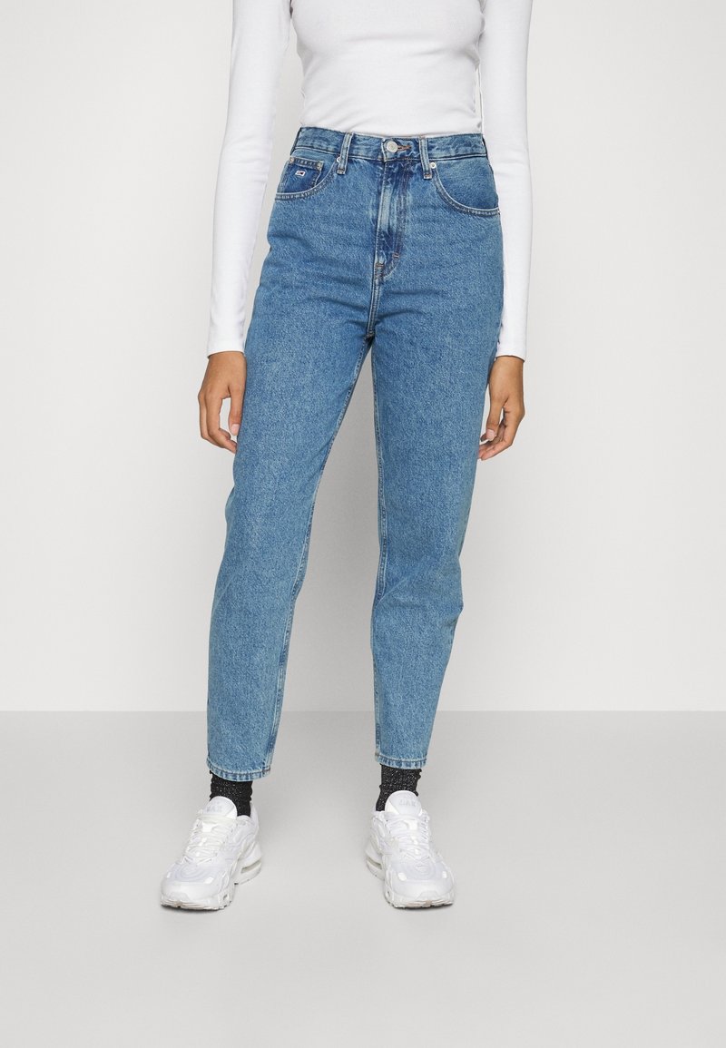 Jeans in denim blu a vita alta con vestibilità dritta, dotati di tasche frontali e chiusura con bottone, abbinati a sneakers bianche.