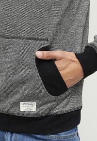 Sudadera con capucha en gris y negro, fabricada con un tejido texturizado, cuenta con un bolsillo frontal y puños acanalados. La etiqueta en la esquina inferior indica la marca.