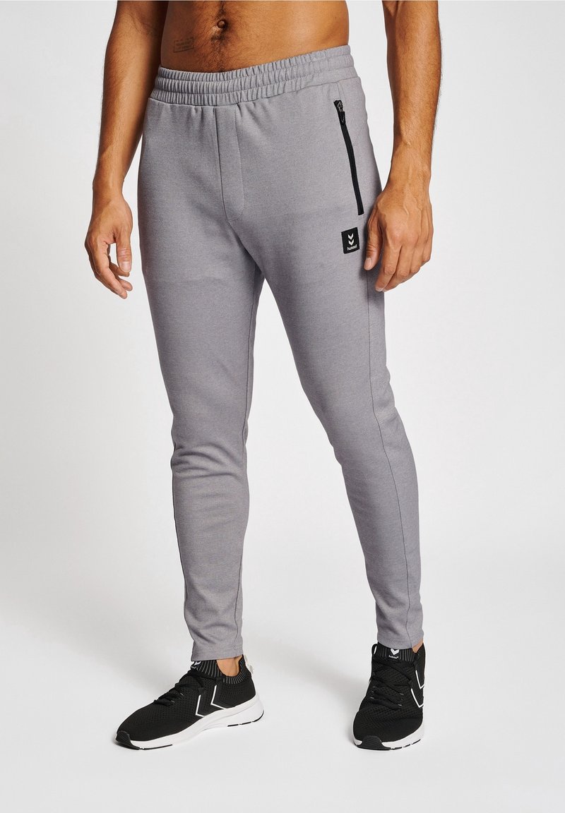 Pantalons de jogging gris, ajustés, avec une taille élastique, une poche zippée sur le côté et un petit logo. Associés à des chaussures de sport noires.