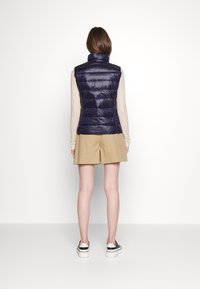 Kamizelka puchowa w głębokim granacie z wysokim kołnierzem, pikowanym wzorem i błyszczącą fakturą, w zestawieniu z beżowymi szortami oraz czarno-białymi trampkami.