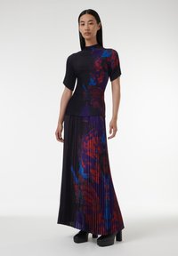 Top de punto negro con canalé y mangas cortas, combinado con una falda larga plisada que presenta un estampado abstracto colorido en tonos rojos y azules. Zapatos de plataforma.