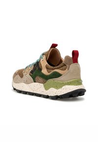 Zapatilla deportiva con paneles de ante beige, verde y marrón, cordones azules, lengüeta trasera roja y suela blanco suculenta moteada, sobre fondo blanco.