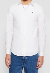 Man i vit skjorta med knappar och en liten logotyp på bröstet, och blå jeans, stående mot en enkel vit bakgrund.