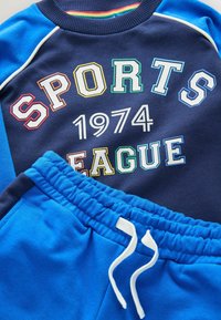Námornícky sveter s nápisom "SPORTS LEAGUE 1974" v viacfarebných písmenách, v kombinácii s modrými teplákmi s elastickým pásom a šnúrkou.